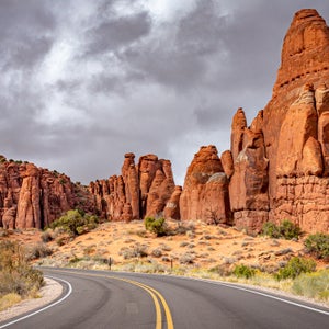 The sun brightly illuminates the red rock formations of the Fiery Furnace area of Arches National Park, contrasted by dark storm clouds in the background. The park road can be seen in the foreground.