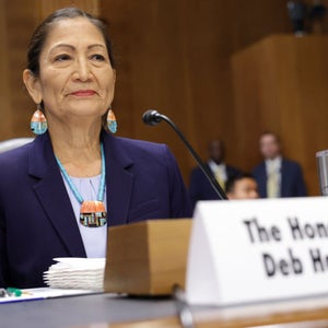 U.S. Interior Secretary Deb Haaland, a woman with dark hair wearing a purple suit jacket, sits behind a microphone while testifying in the Senate Office Building.