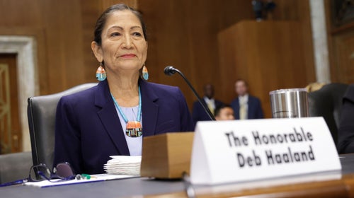 U.S. Interior Secretary Deb Haaland, a woman with dark hair wearing a purple suit jacket, sits behind a microphone while testifying in the Senate Office Building.