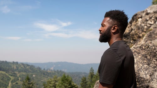 Baratunde Thurston stands on a cliff and looks out over a mountainous, forested landscape.