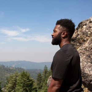 Baratunde Thurston stands on a cliff and looks out over a mountainous, forested landscape.