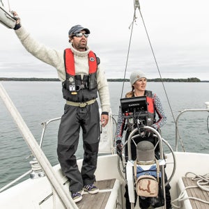 A man an a woman are on the deck of a boat. She is at the helm, he is standing. They both look faintly puzzled.