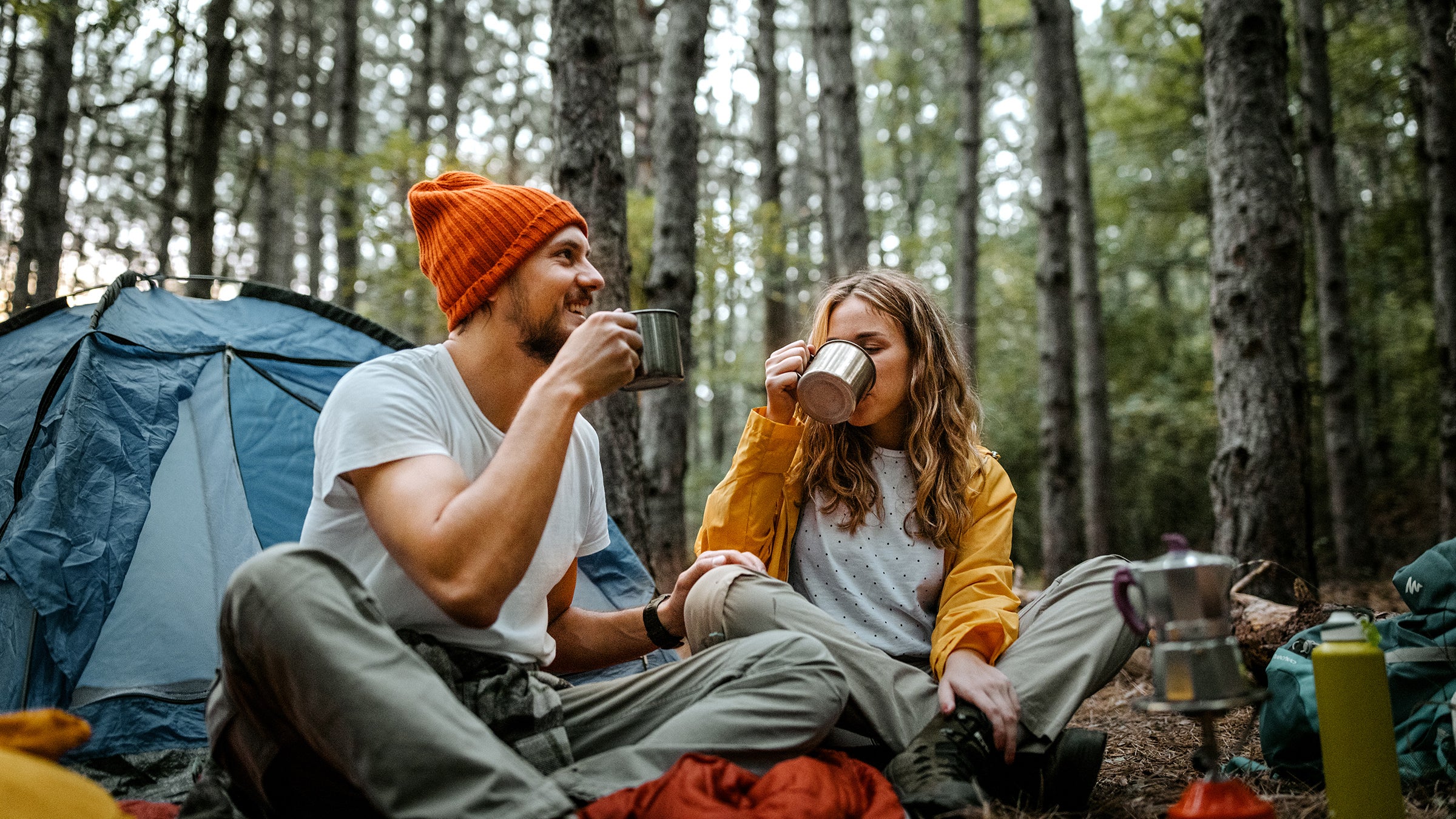 Low angle view of young male and female friends sitting on blanket on ground outside tent during hiking and drinking coffee