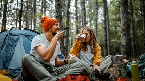 Low angle view of young male and female friends sitting on blanket on ground outside tent during hiking and drinking coffee