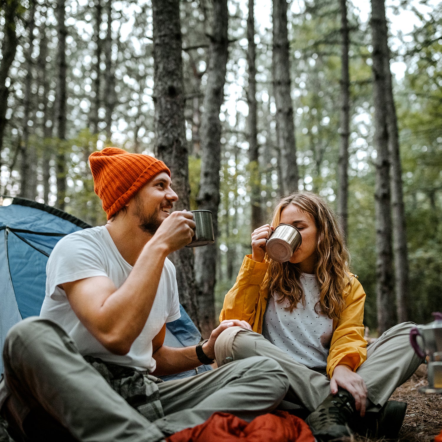 Low angle view of young male and female friends sitting on blanket on ground outside tent during hiking and drinking coffee