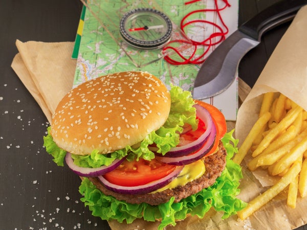 A juicy hamburger and fries sit on a table with a map, compass, and pocket knife.