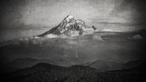 A black and white photo of Mt. Hood. There are smaller mountains in the foreground and clouds around the base of Mt. Hood. The image looks old, faded, and ominous.
