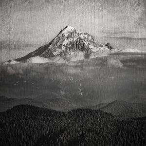 A black and white photo of Mt. Hood. There are smaller mountains in the foreground and clouds around the base of Mt. Hood. The image looks old, faded, and ominous.
