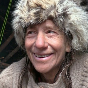 A contestant on Alone wears a fur hat inside a structure and smiles.