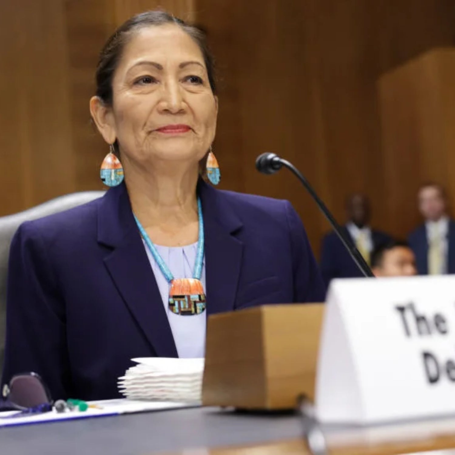 U.S. Interior Secretary Deb Haaland, a woman with dark hair wearing a purple suit jacket, sits behind a microphone while testifying in the Senate Office Building.