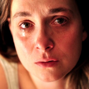 A white woman with blonde hair looks directly at the camera, crying quietly. She is wearing a white tank top.