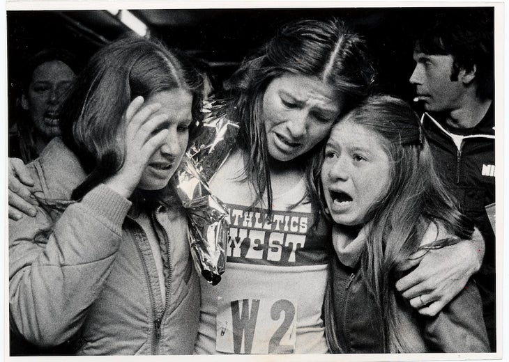 Black and white photo from the finish of the Boston Marathon, where Patti Dillon is held by two sisters at the finish.