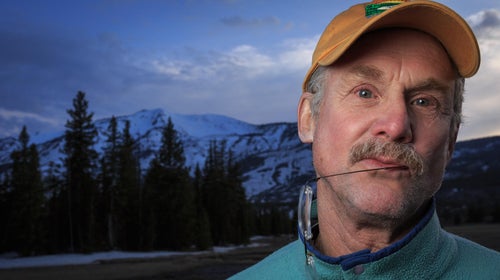 David Quammen is an older white man with a gray mustache. He is standing in front of a snowy mountain range wearing an orange baseball cap. He has his glasses hanging from his mouth and looks pensive.