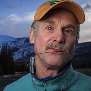 David Quammen is an older white man with a gray mustache. He is standing in front of a snowy mountain range wearing an orange baseball cap. He has his glasses hanging from his mouth and looks pensive.