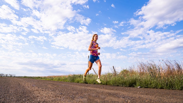 a woman in a pink singlet and blue shorts runs on a dirt road with a blue sky in the background