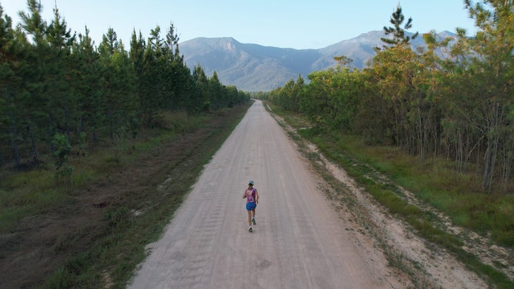 a female runner follows a long dirt road in Australia, as seen from a drone above.