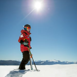 A ski patroller in a red coat stands on a snow-covered ridge, with mountains lining the horizon behind them. They are holding their skis and poles.