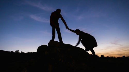 A silhouetted person helps another onto a rock
