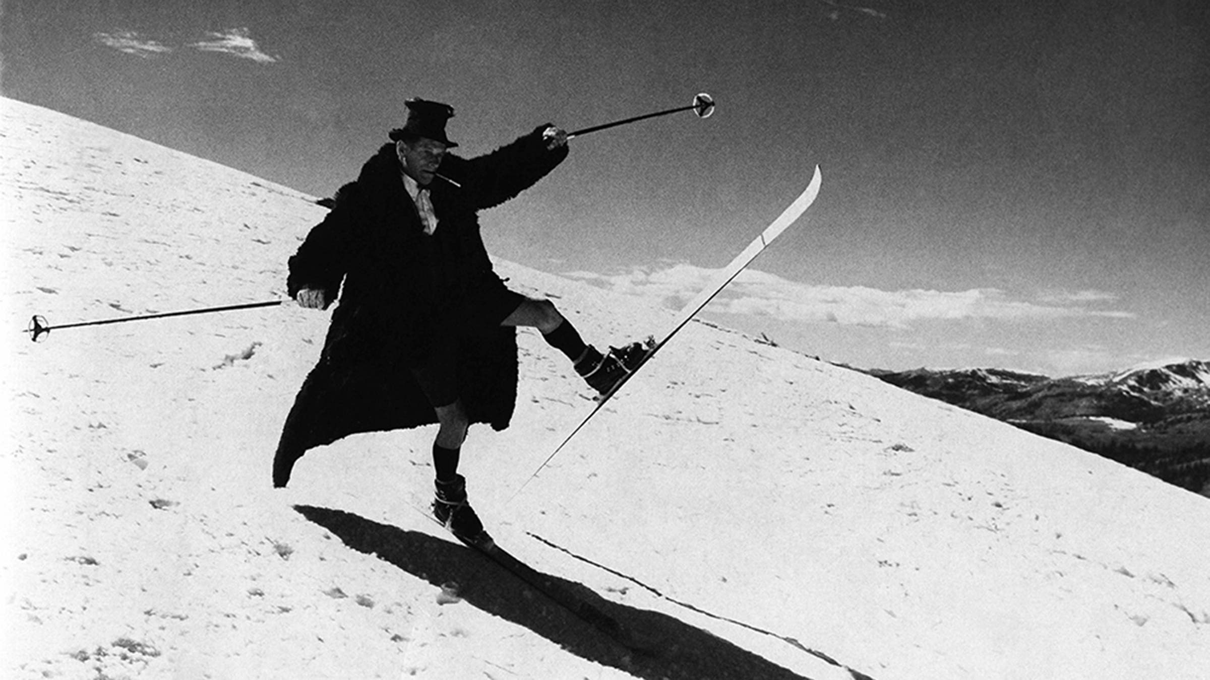 Ralph Jackson skiing an early-season run on Independence with his traditional hat, fur coat, cigarette holder in mouth, and shorts