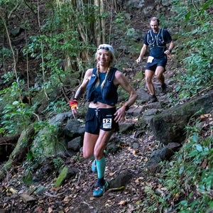 A woman and her pacer run in Hawaii on rooted trail. They are smiling and the forest is green.