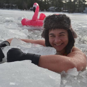 A smiling woman wearing a fur hat and gloves is immersed in icy water. She hugs a chunk of ice to her chest as an inflatable flamingo looks on.
