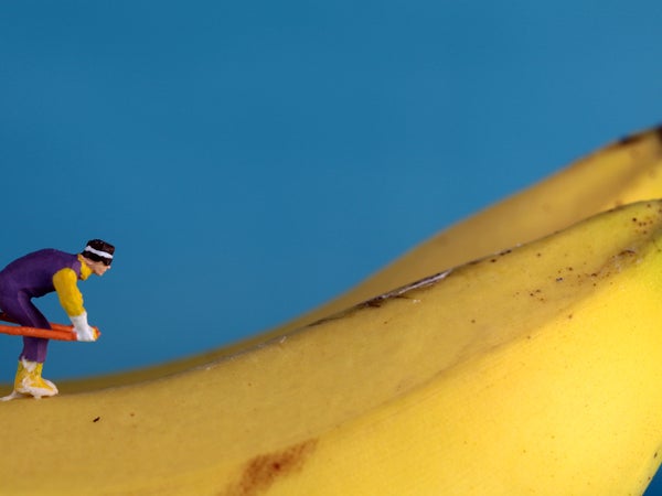 A one-centimeter figurine of a ski jumper looks ready to sail off the end of a banana, against a solid blue background.