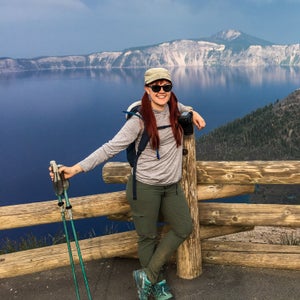 Emily (white woman with red hair, wearing hiking clothes and holding trekking poles) stands in front of Crater Lake, with mountains in the background.