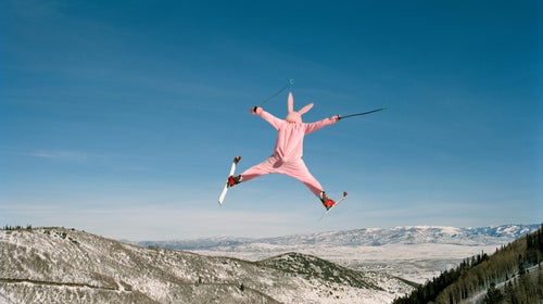 A person in a pink bunny suit on skis flies through the air, spread-eagled.