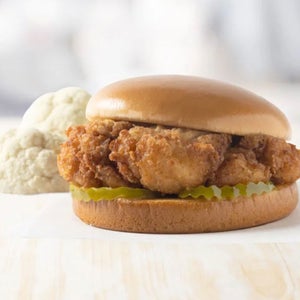 A Chick-fil-A fried cauliflower sandwich--a browned piece of cauliflower on a shiny bun with pickle slices rests on a white napkin. A chunk of fresh cauliflower is in the background.