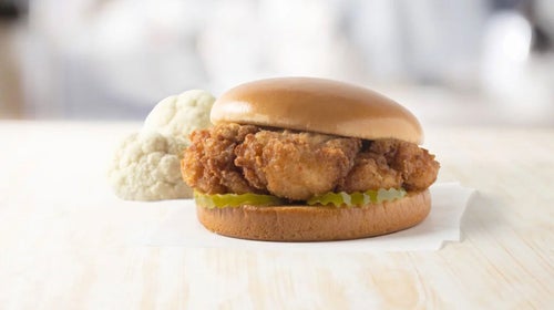 A Chick-fil-A fried cauliflower sandwich--a browned piece of cauliflower on a shiny bun with pickle slices rests on a white napkin. A chunk of fresh cauliflower is in the background.