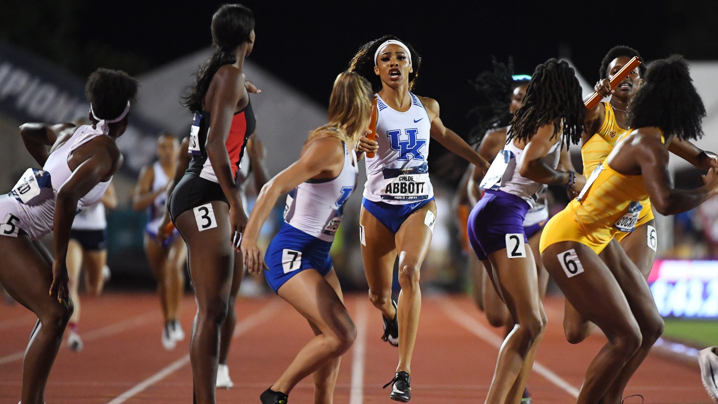 A lineup of collegiate track athletes wait for the baton at a relay.