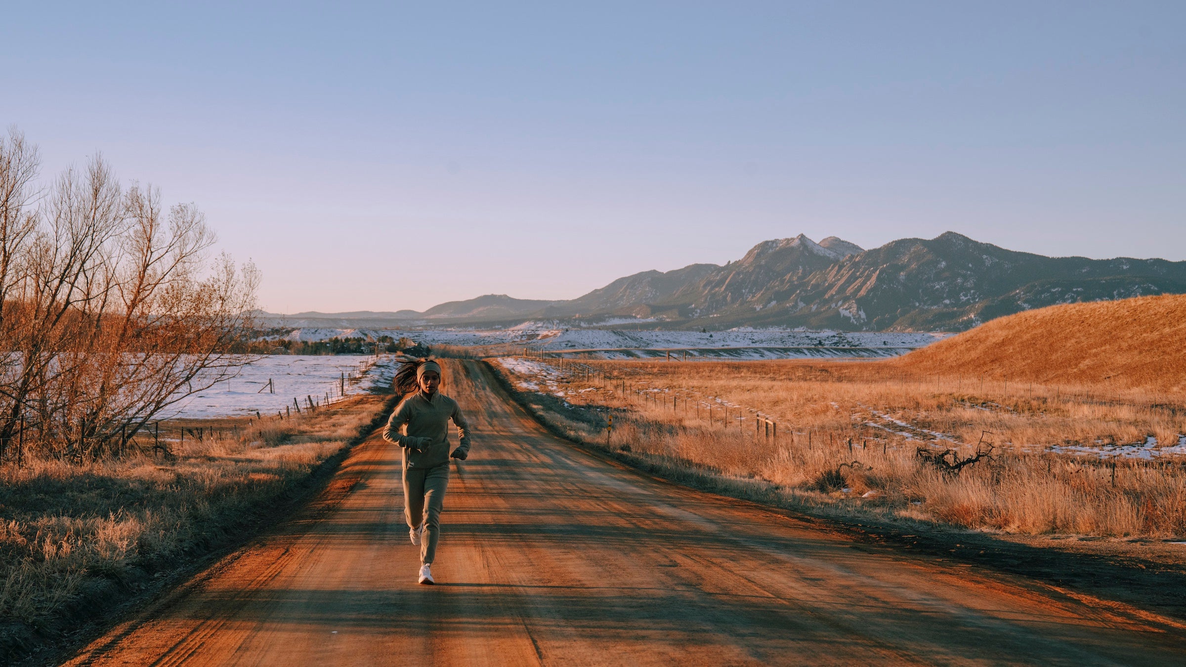 a woman dressed in grey run along a diry road that is brown and orange. There is snow and mountains.