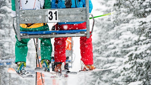 Two people sit on a ski lift while it snows. They are seen from behind, and their faces are hidden.