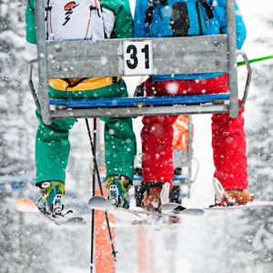 Two people sit on a ski lift while it snows. They are seen from behind, and their faces are hidden.