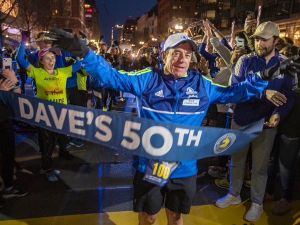 Dave McGillivray runs through a blue finish line tape surrounded by a large crowd