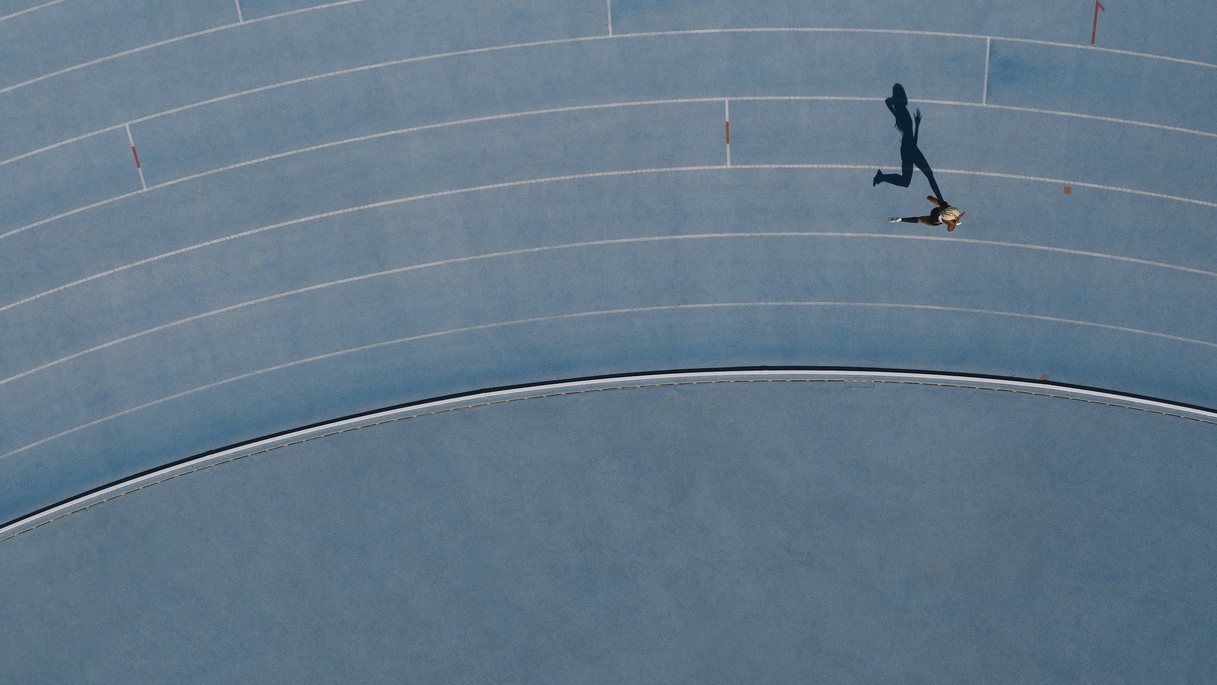 A woman runs around the bend of a light blue track, her shadow cast