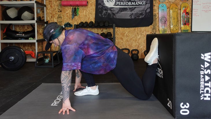 One of the mobility exercises for knee pain. A woman in a half-kneeling position bends her back leg so that her shin is parallel with a box. She is leaning forward with her hands on the floor.