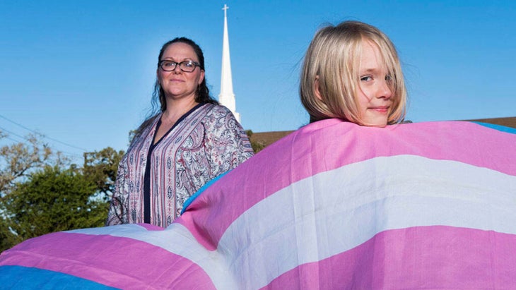 A blonde girl draped in a light blue, light pink, and white transgender flag poses in front of a woman outdoors. The steeple of a church is seen in the background.