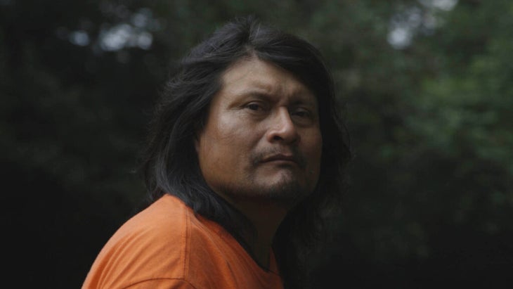 A portrait of a Rama man in front of trees. He has long dark hair and is wearing an orange t-shirt.
