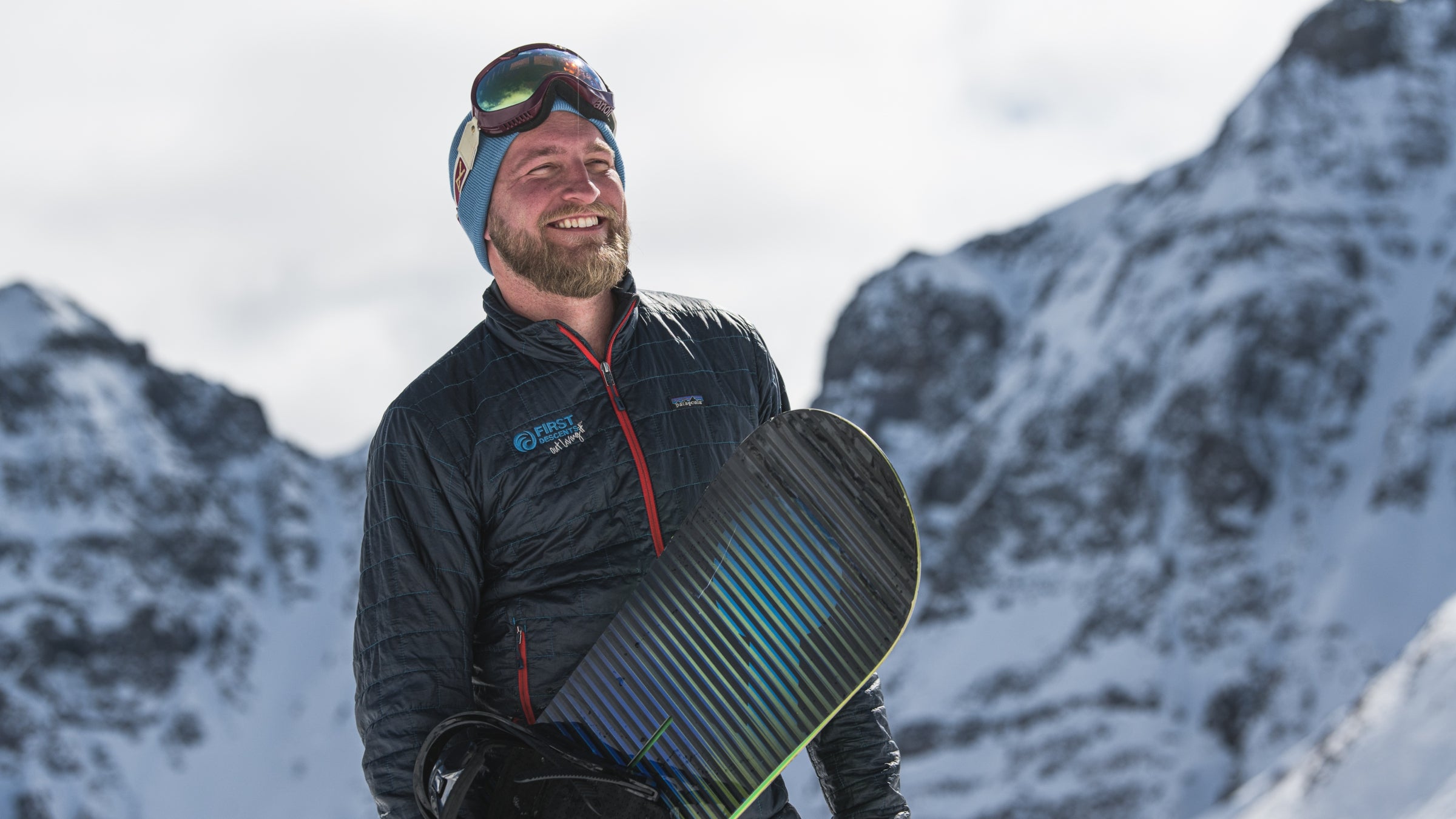 Conor Hall (a white man with a beard) holds a snowboard and looks into the distance with snowy peaks in the background.