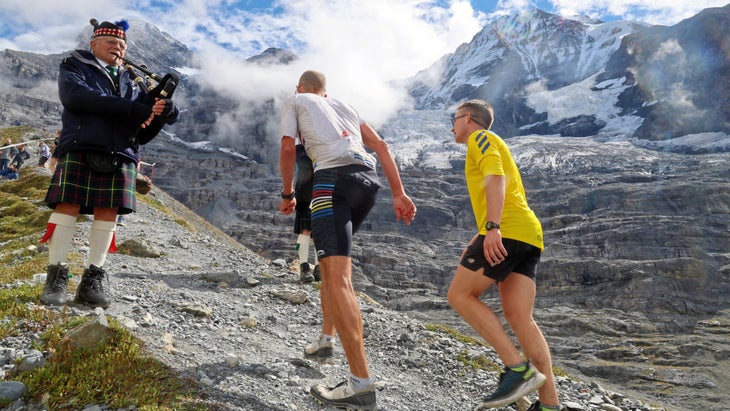 Two men hike up a steep tail while an older man plays music for them