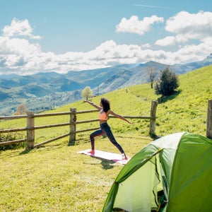 Black female practicing Crescent lunge on mat having yoga session on green meadow with tent against mountain valley