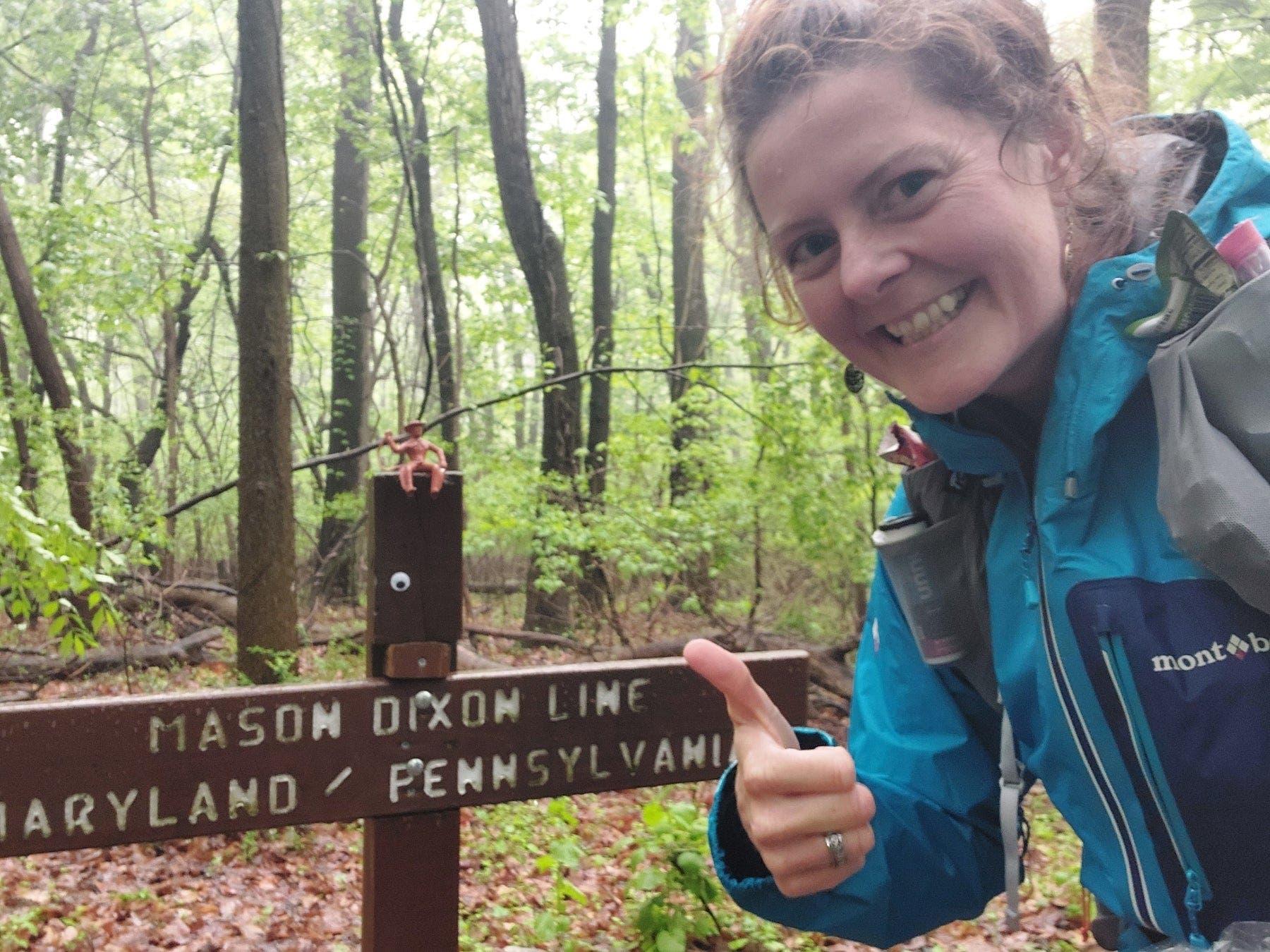 Heather Anderson smiles with a thumbs up in front of a Mason Dixon line sign on the trail.