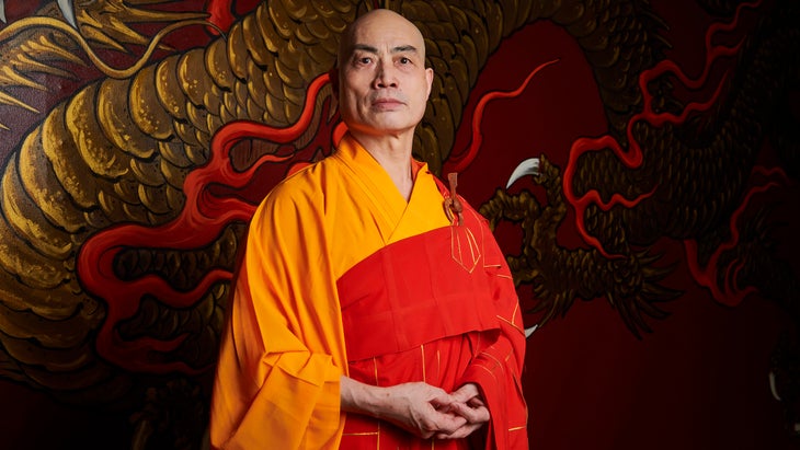 A monk in an orange and red robe stands in front of a dragon painting