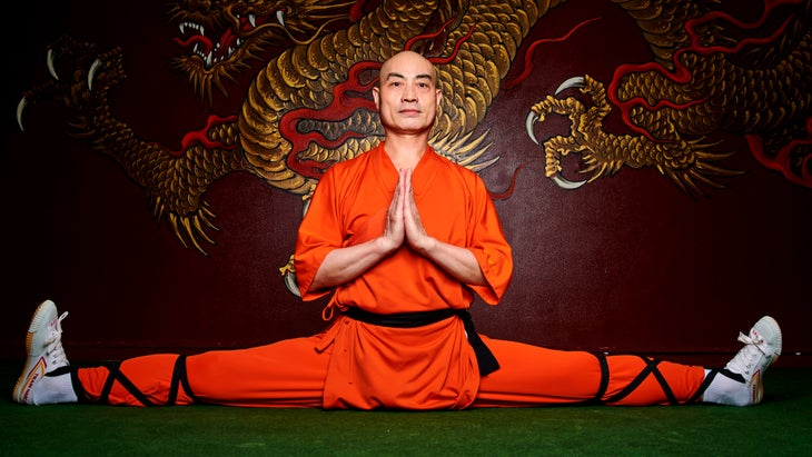Monk in an orange suit does the splits on a green mat.