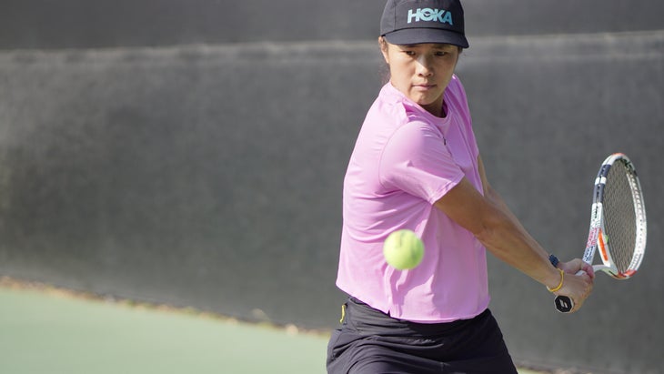 A woman in a purple shirt and black hat swing a tennis racquet