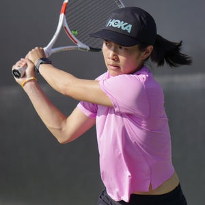 A woman in a purple shirt and black hat swing a tennis racquet
