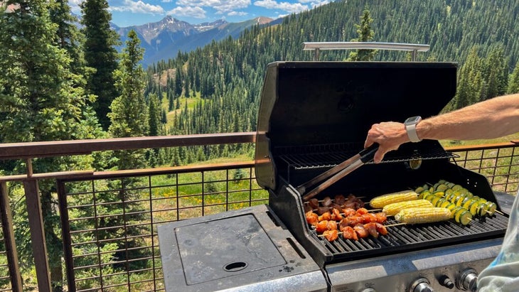 A person grilling corn, zucchini skewers and meat, with an incredible mountain view off the porch.