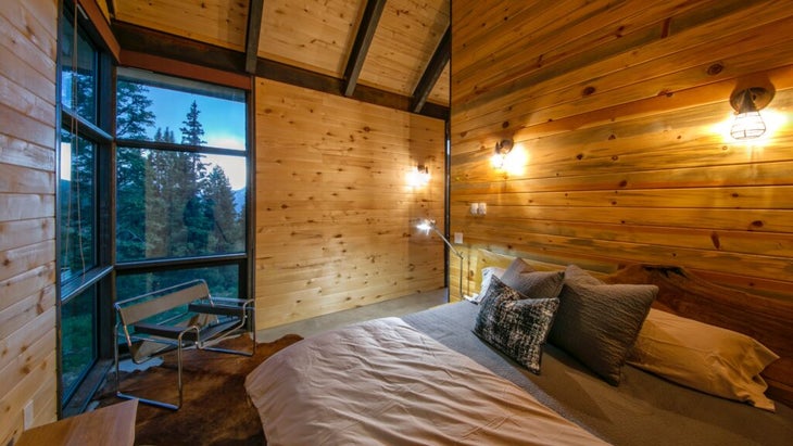 The king bedroom of the Thelma Hut, with wood-paneled walls and two windows that look out at a pine forest
