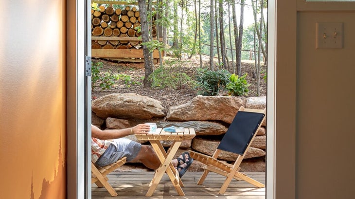 The back porch of a cabin, with two wooden folding chairs and a folding table, with the woods in the background